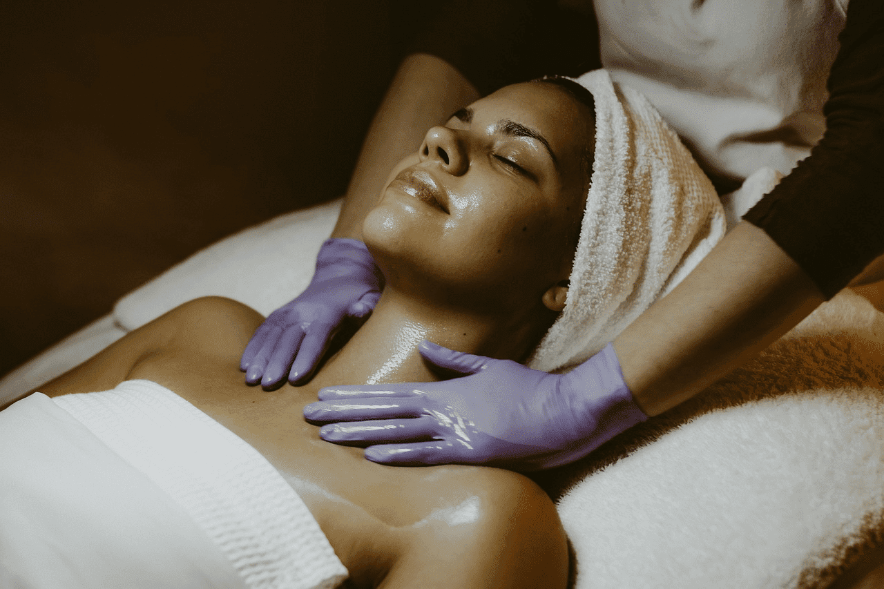 Woman enjoying a spa facial massage, wearing a towel and attended by gloved hands.