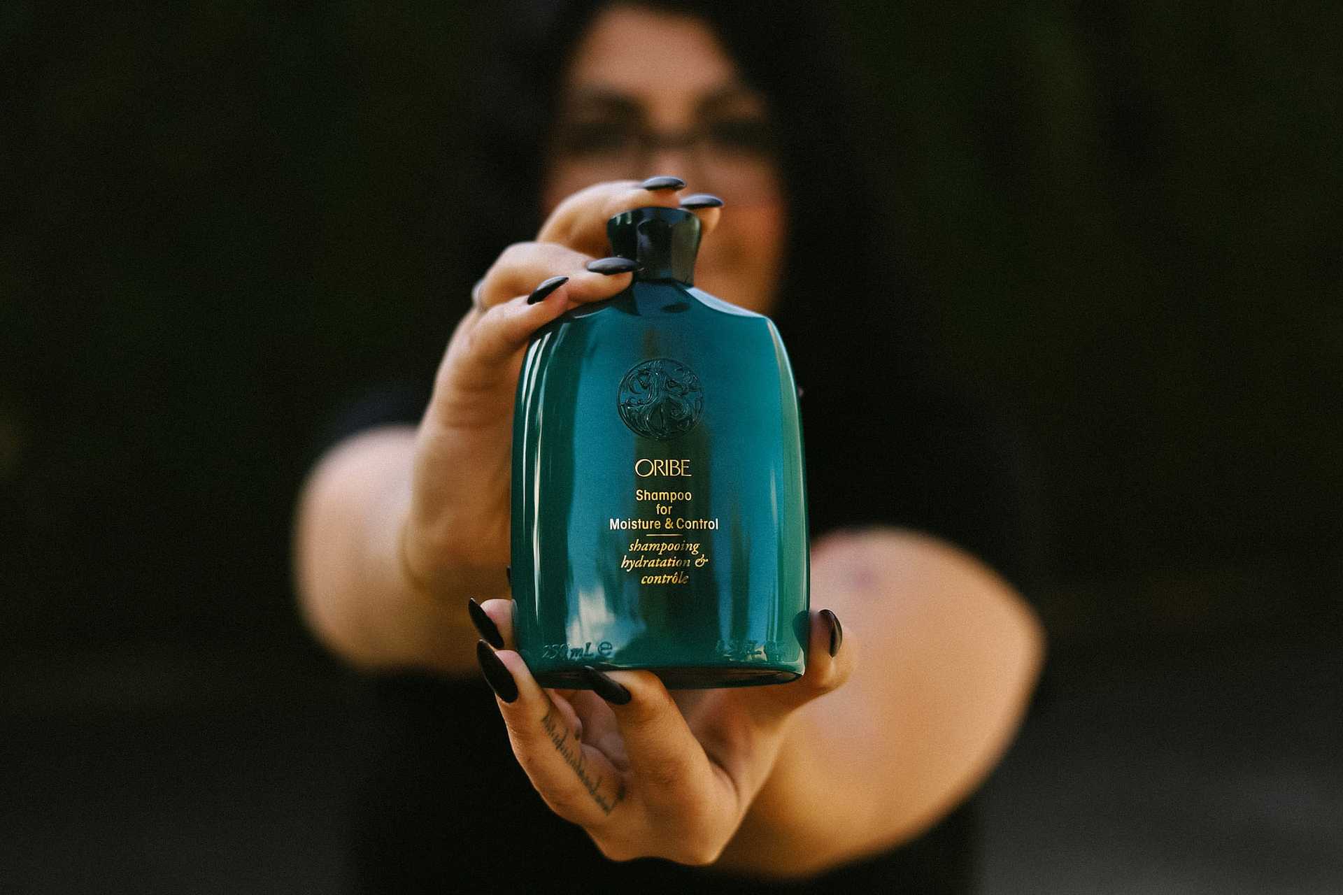 Person holding a green Oribe shampoo bottle towards the camera.