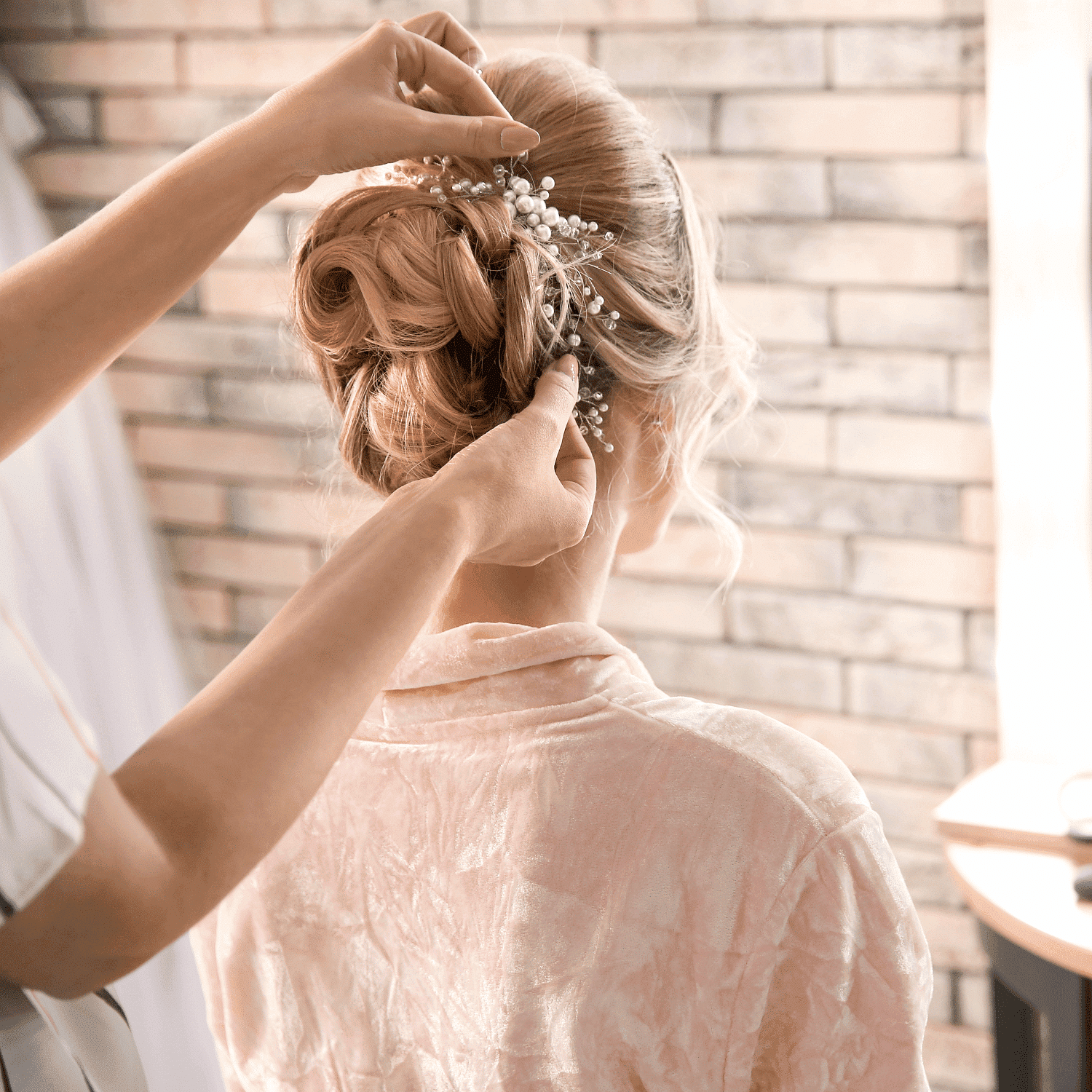 Hairstylist arranging elegant updo with decorative hairpin on woman in pink robe.
