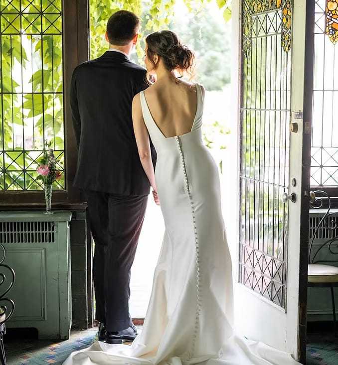 Bride and groom standing in doorway, elegant white gown, natural light filtering through leaves.