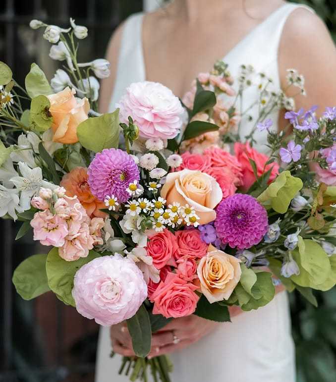 Bride holding a vibrant bouquet of pink, purple, and orange flowers against a blurred background.