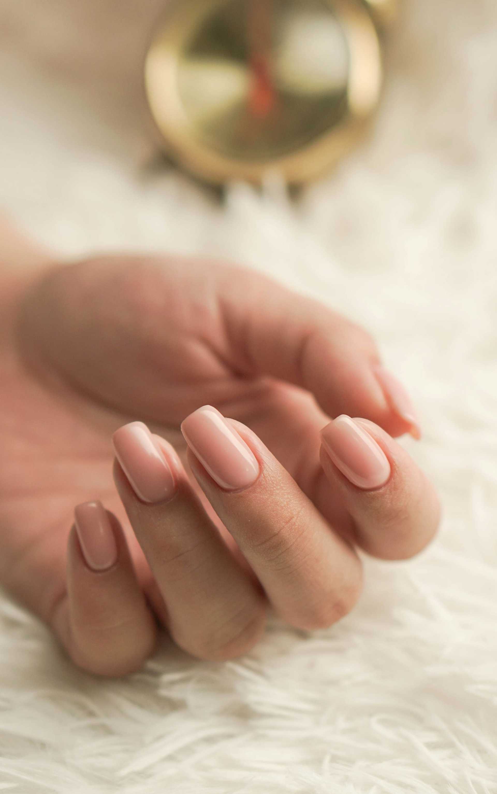 A hand with manicured nails rests on a soft white surface, featuring a blurred gold accessory.