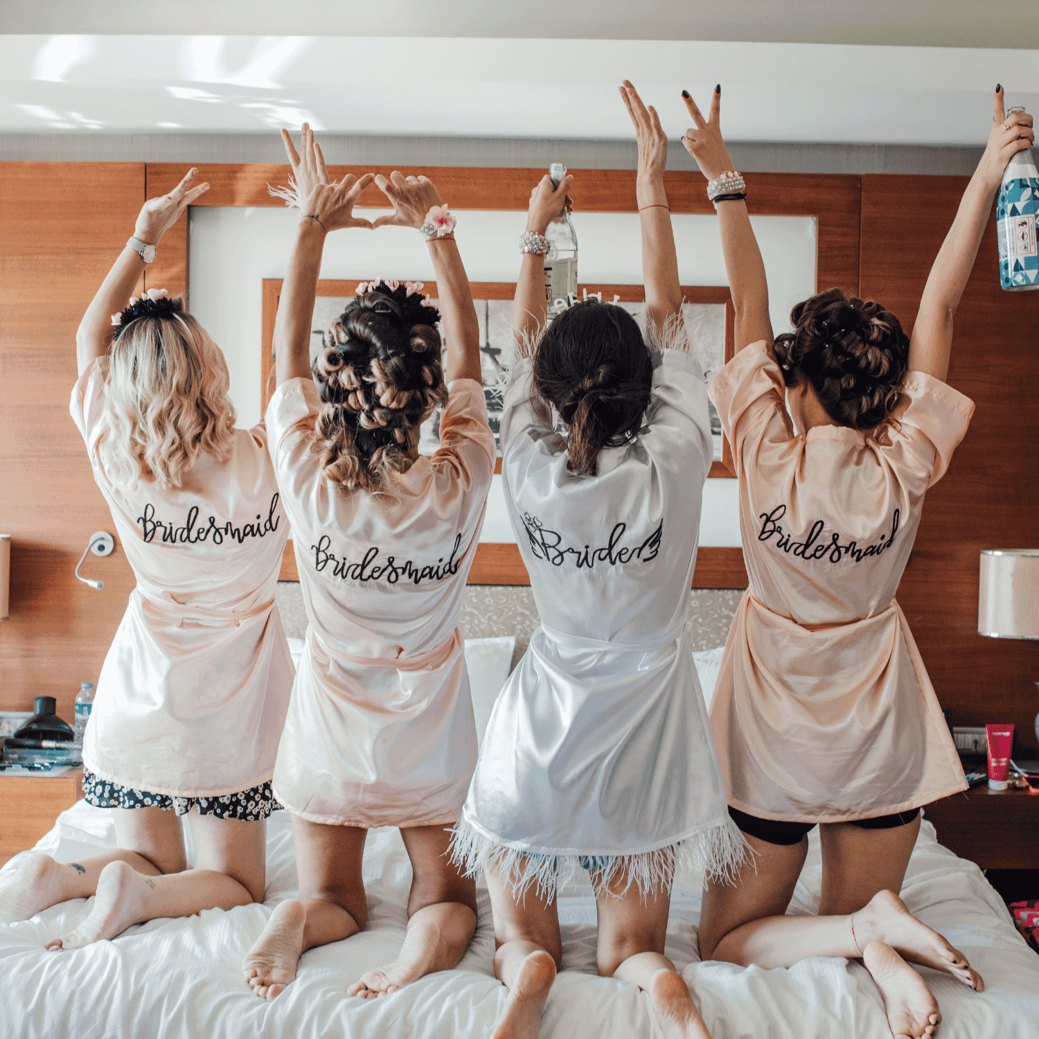 Bridesmaids and bride celebrating with raised arms on a bed in matching robes.