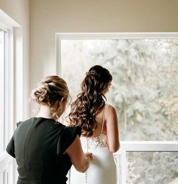 Bride getting ready by a window with assistance from another woman.