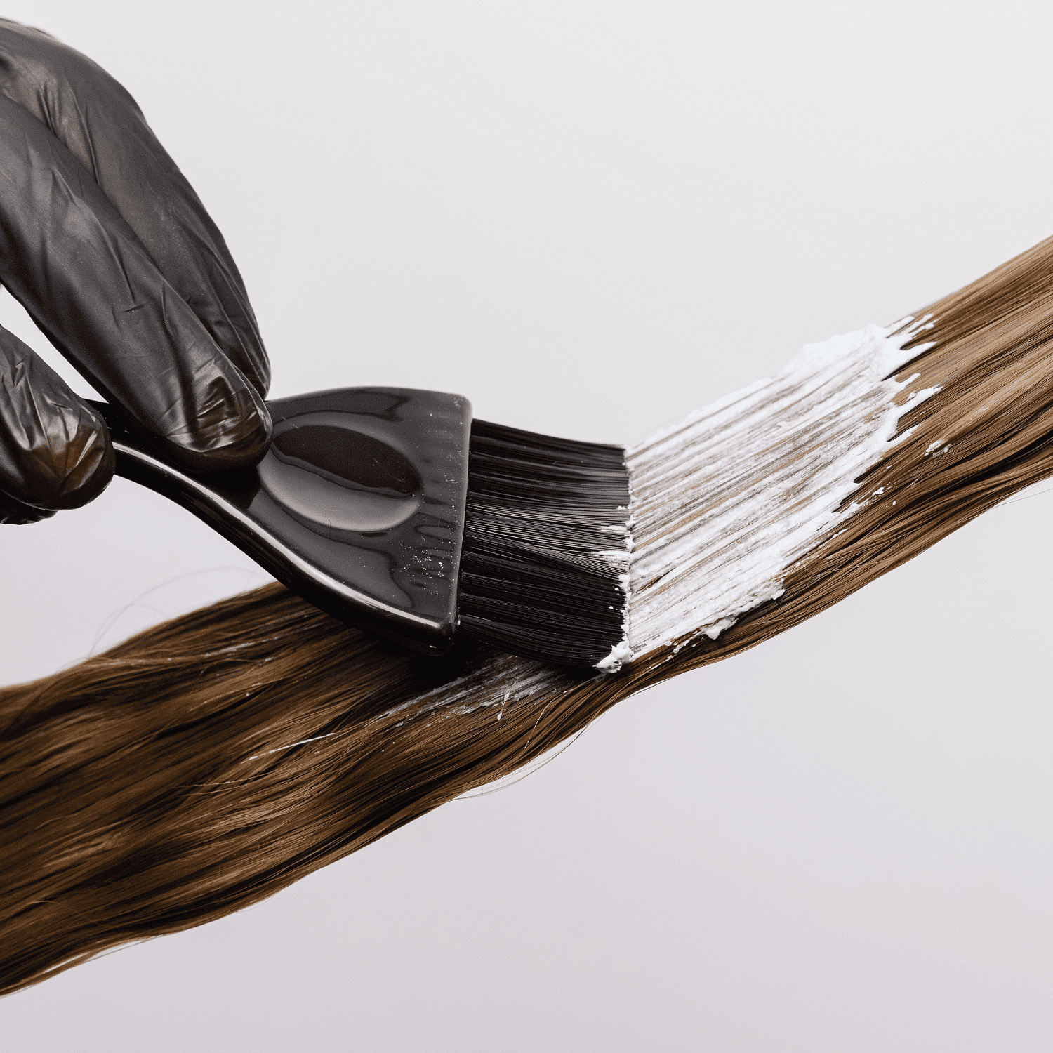 Hair being dyed with a brush, black gloves, and white dye on brown strands.