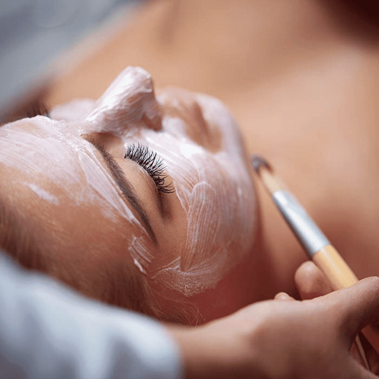 Person receiving a facial treatment with a brush applying a white mask to their face.