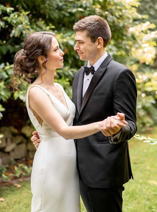 Couple in elegant attire dancing together outdoors, surrounded by greenery.