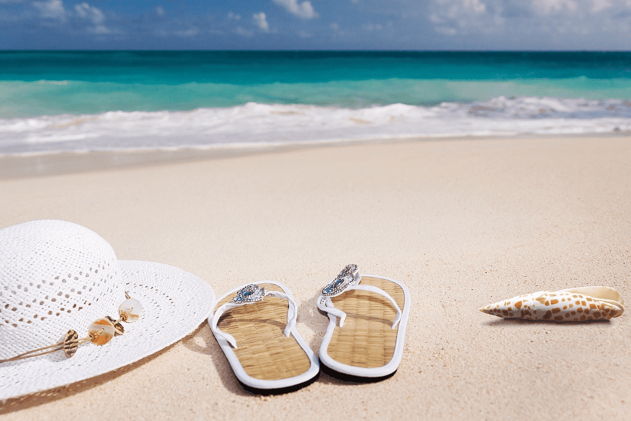 Beach scene with flip-flops, sun hat, and seashell on sandy shore.